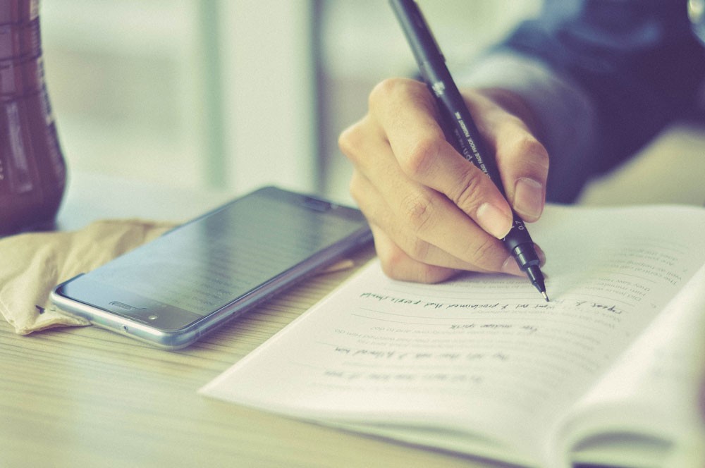 mano escribiendo encima de una libreta y al lado un teléfono 